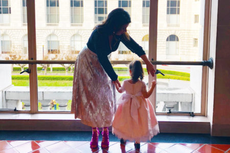 A woman holds her young daughter's hands as they stand near an expansive window during the day