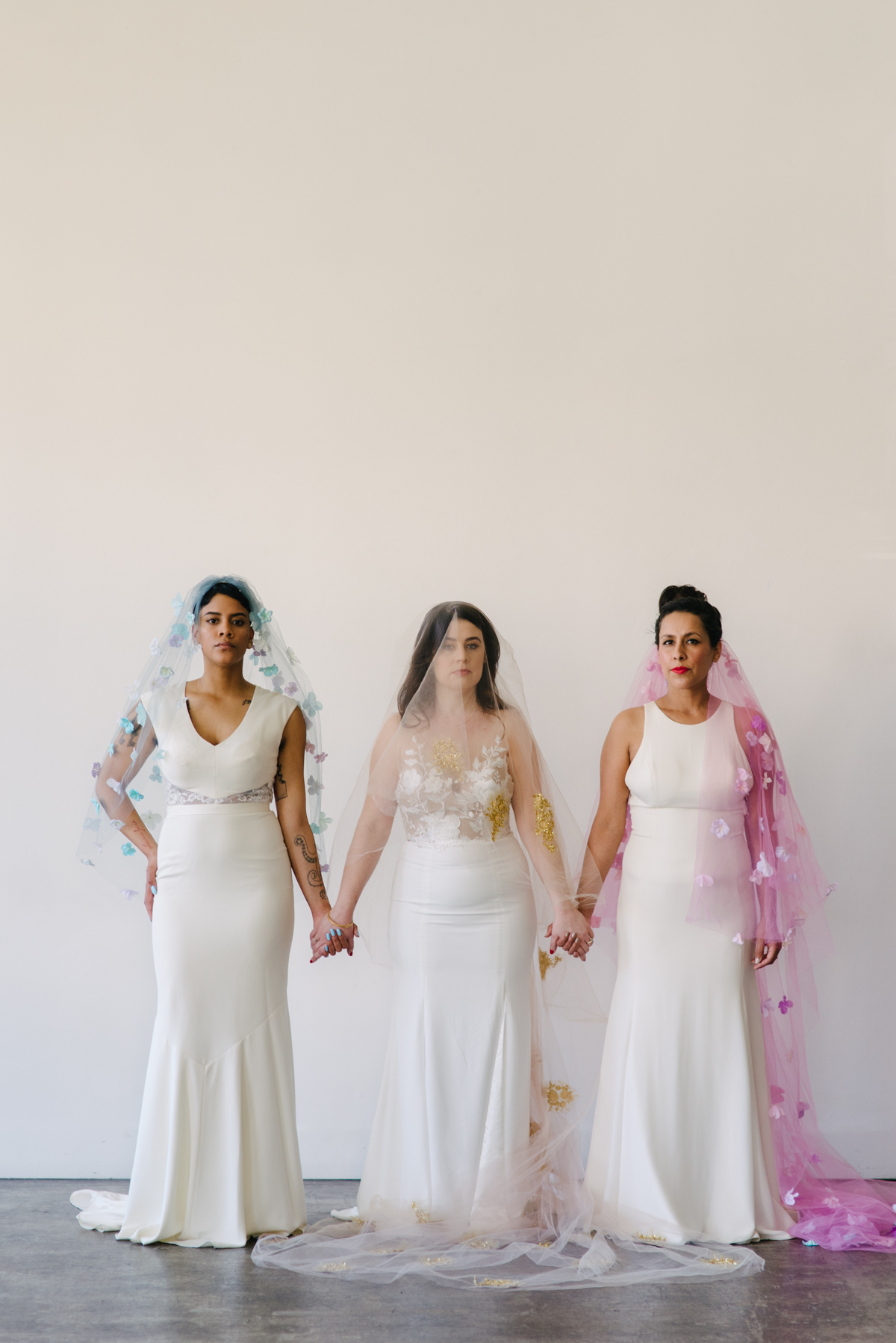 Three women stand side by side holding hands while wearing different color wedding veils