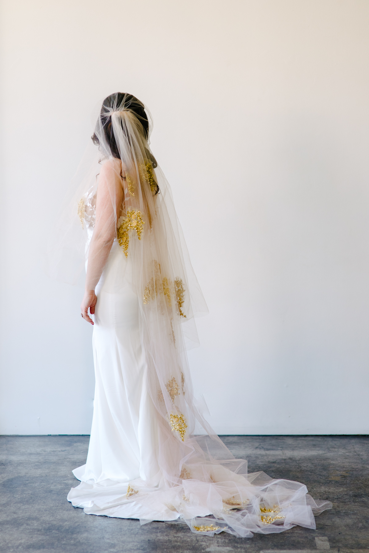 A full body reverse view of a woman wearing a long wedding veil with gold accents