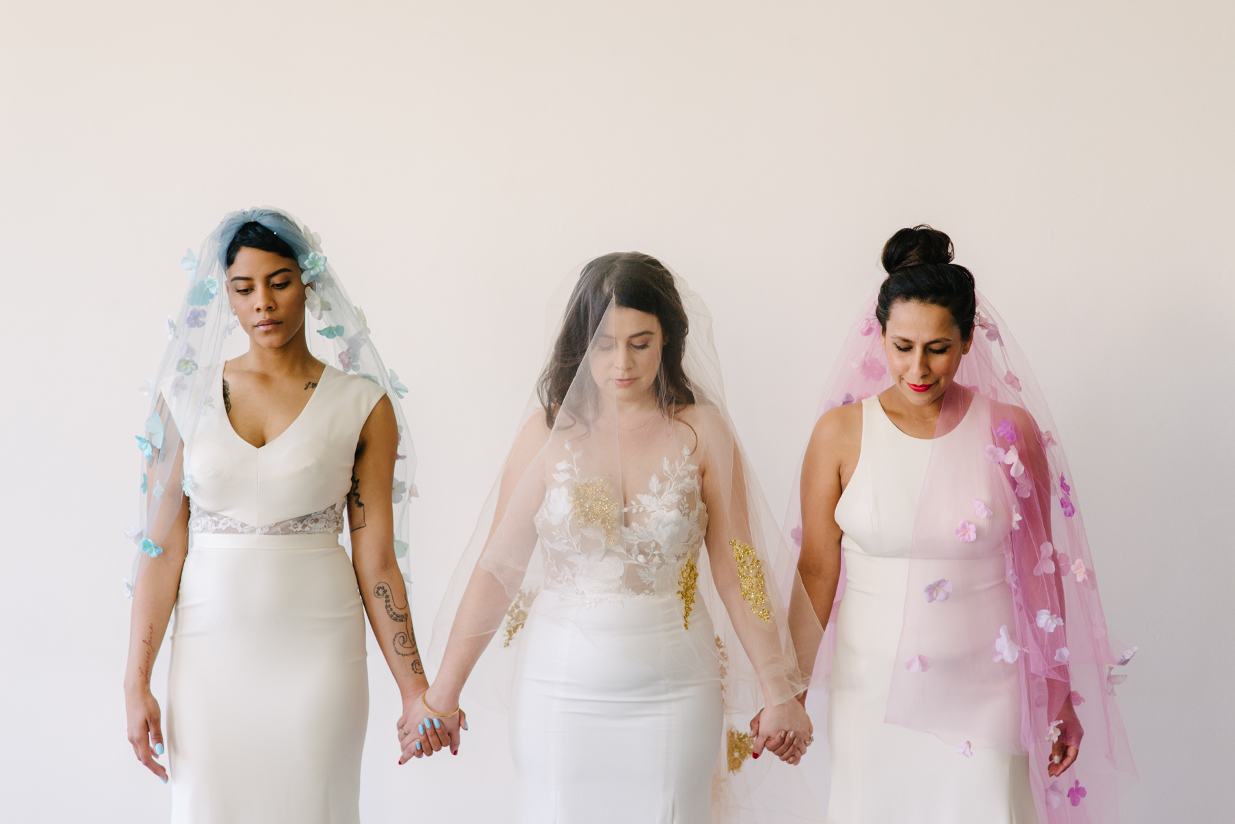 Three women hold hands in a moment of silence while wearing different colored wedding veils