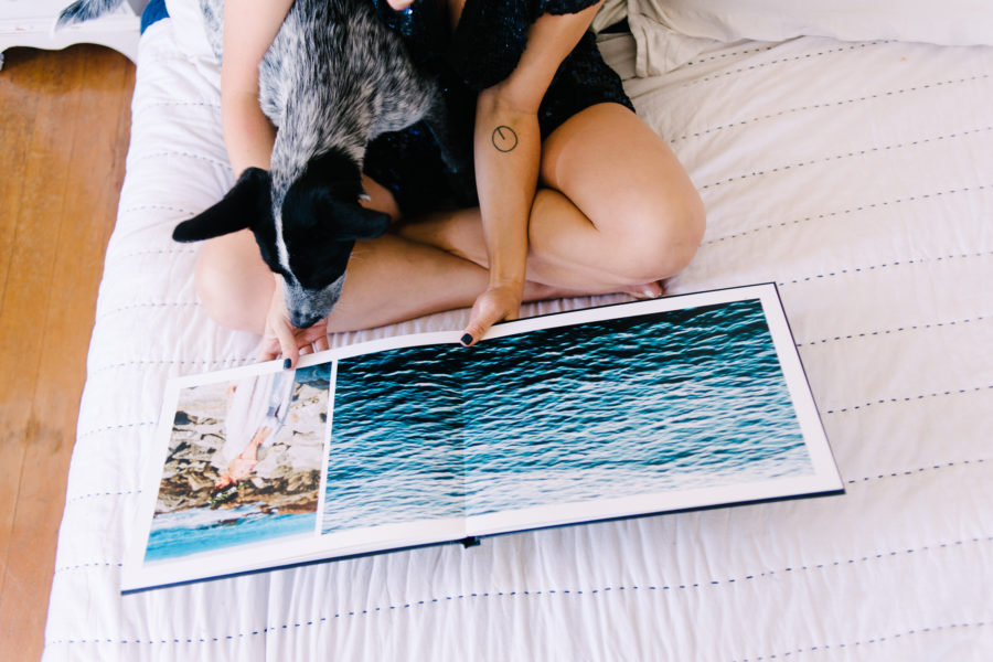 A person sits on a bed looking through a photo book while holding a black and white dog on their lap