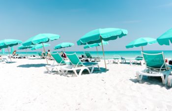 Teal beach umbrellas and lounge chairs on a white sand beach, overlooking crystal teal ocean and blue sky