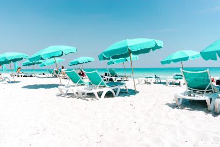 Teal beach umbrellas and lounge chairs on a white sand beach, overlooking crystal teal ocean and blue sky