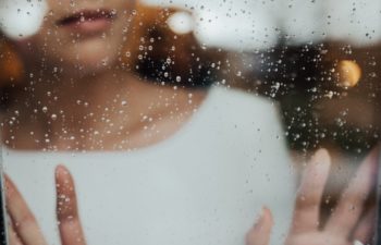 A woman in white stands on the other side of a rainy window, hands pressed against the glass, looking out