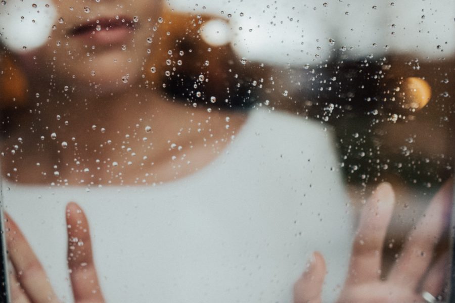 A woman in white stands on the other side of a rainy window, hands pressed against the glass, looking out