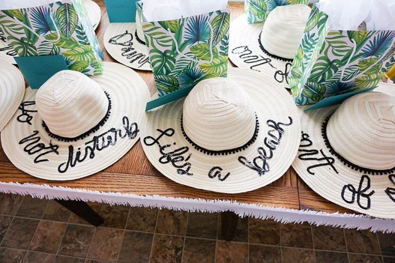 Sun hats with the words "life's a beach" on them, on a table with tropical-themed favor bags for bridal shower favors