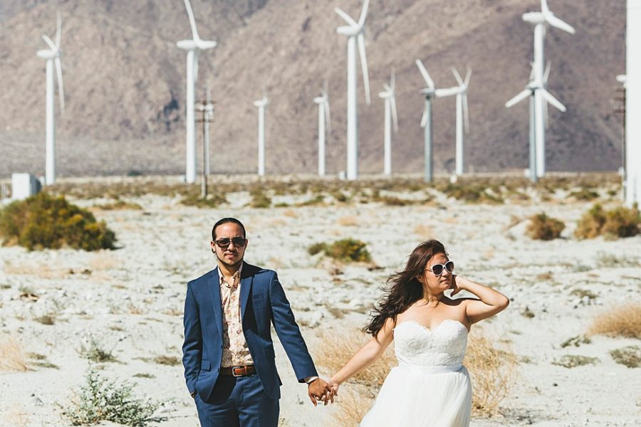 A wedding couple stands just outside a field of white windmills in the desert