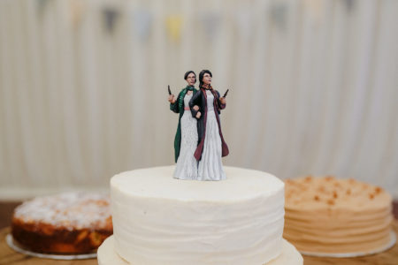 Two womxn in white wedding dresses as cake toppers, one wearing Gryffindor wizarding robes and scarf, the other wearing the same in Slytherin colors. Both hold wands.