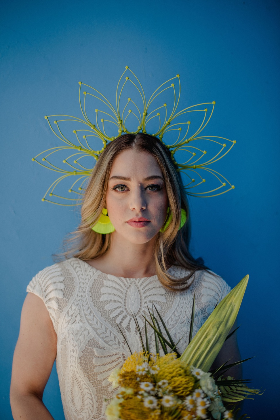 a closeup of a bride wearing an intricate lace gown from allure modest and a neon yellow ziptie headpiece with neon fringe earrings and a bright yellow bouquet. She stands against a blue wall. 