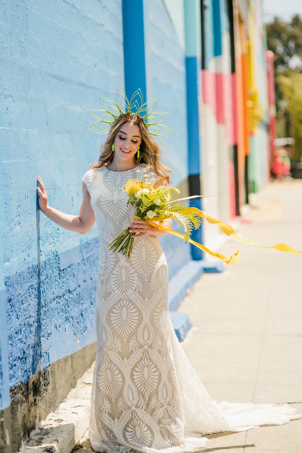 a model stands against a mural holding a large yellow bouquet wearing an allure modest gown and a yellow zip tie headpiece