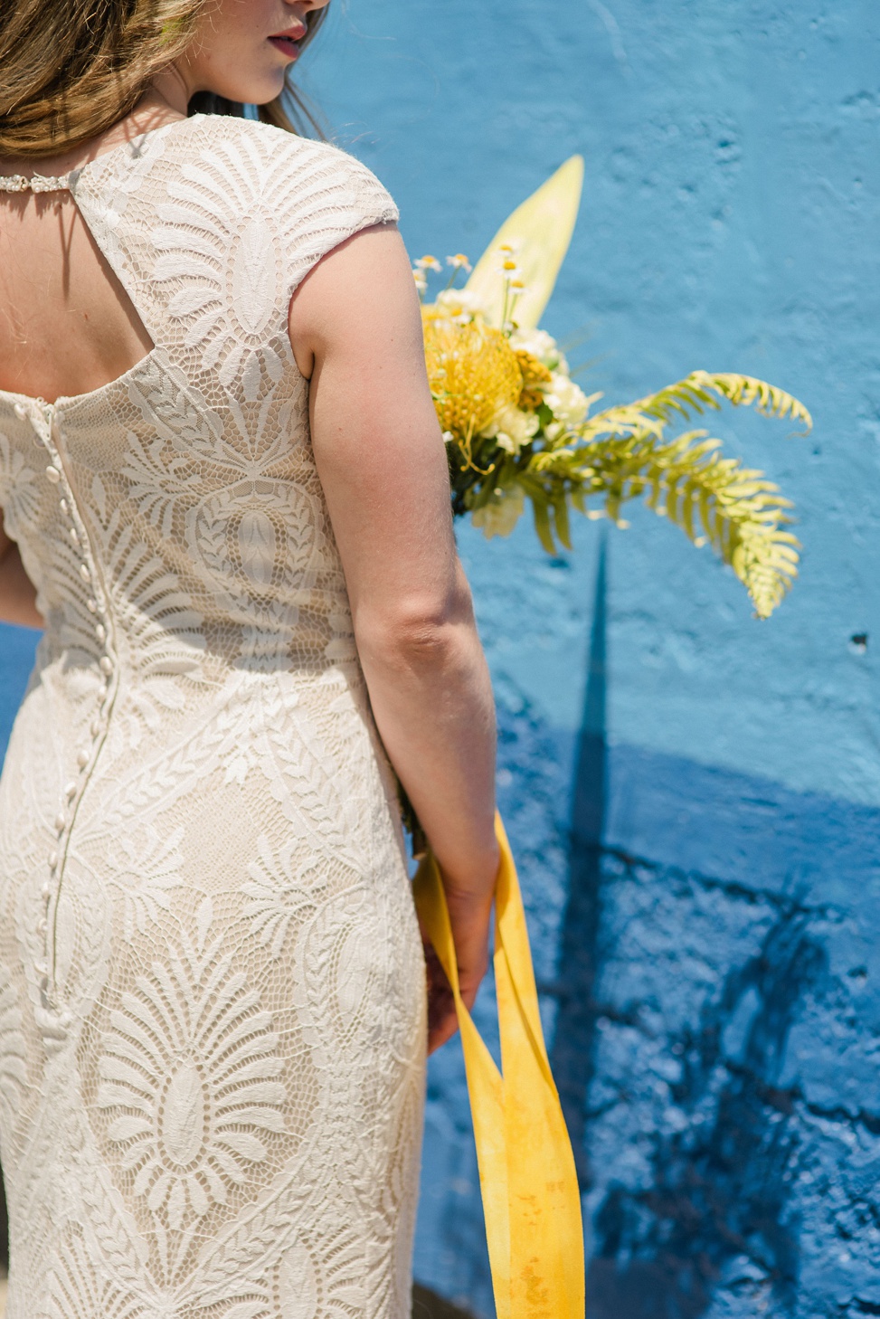 a closeup of the back of a woman wearing a detailed lace gown from allure modest standing against a blue wall holding a large yellow bouquet