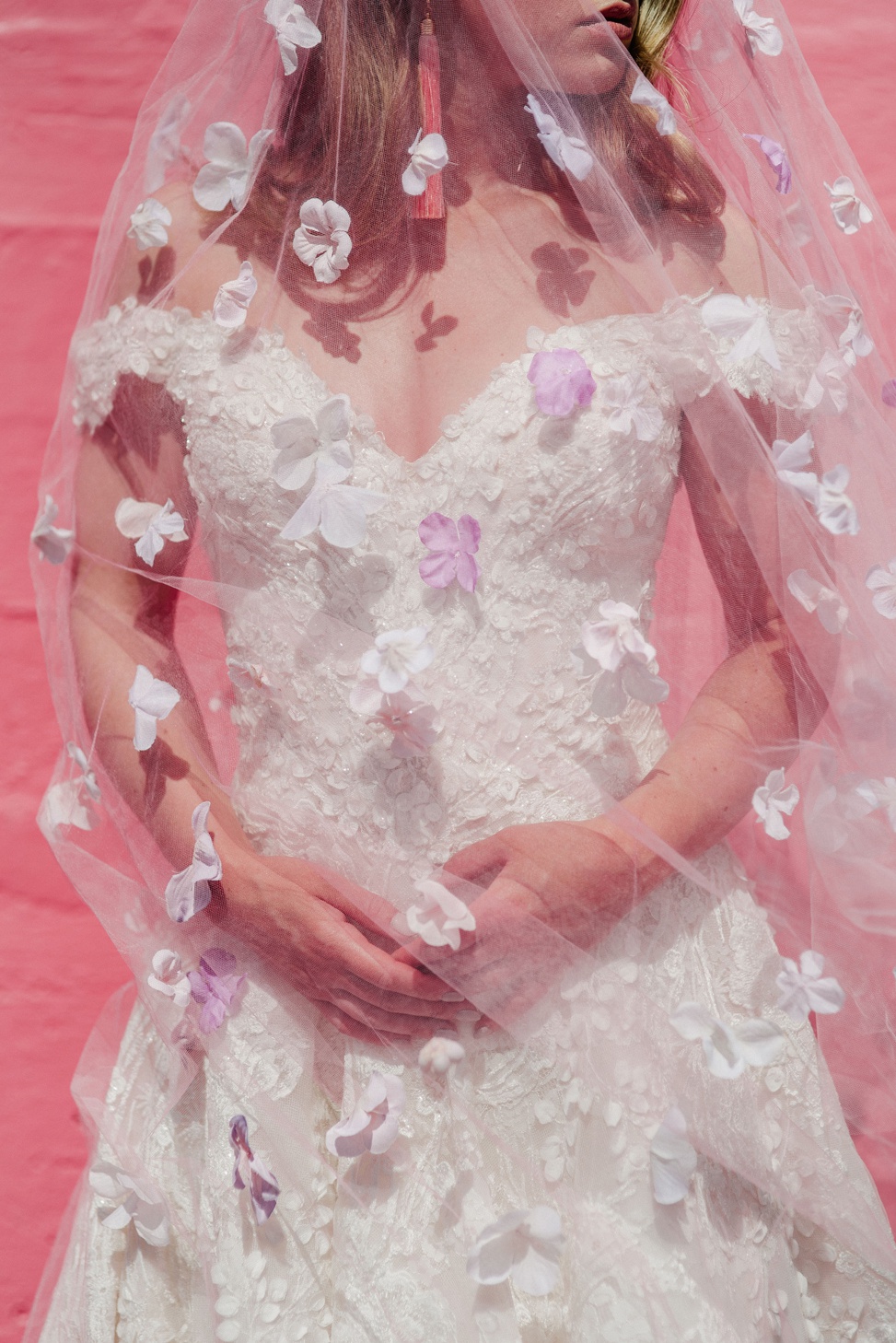 A woman wears allure couture style c520 while standing against a pink wall wearing a pink veil with floral appliques