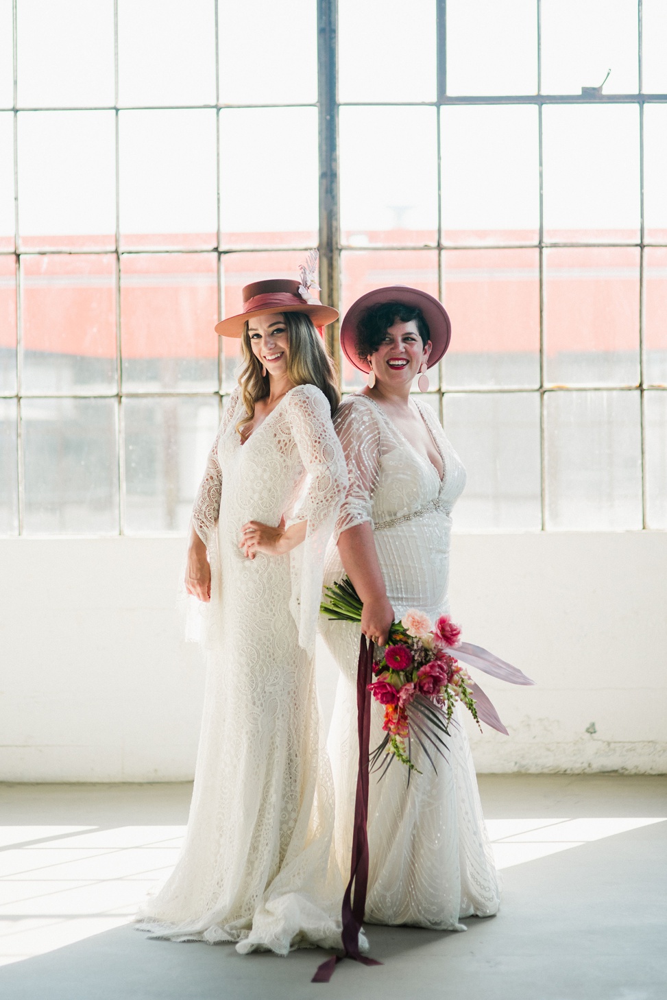 two brides wear intricate long sleeve wedding dresses from madison james in a white warehouse. They each wear a pink brimmed hat