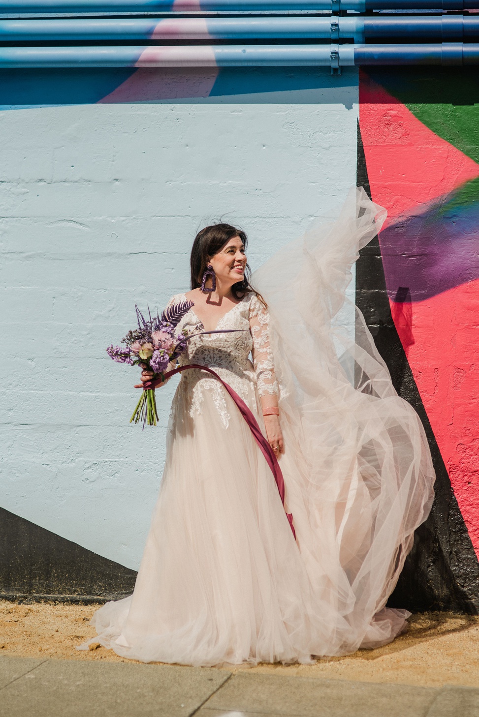 a woman wears a long sleeve lace and tulle gown from madison james while standing against a brightly colored mural. Her dress train is floating in the wind and she holds a purple bouquet.