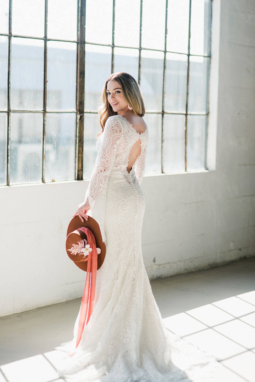 a bride stands in a white industrial building wearing a long sleeve lace gown from madison james. She holds a pink wide brimmed hat with flowers and ribbons cascading