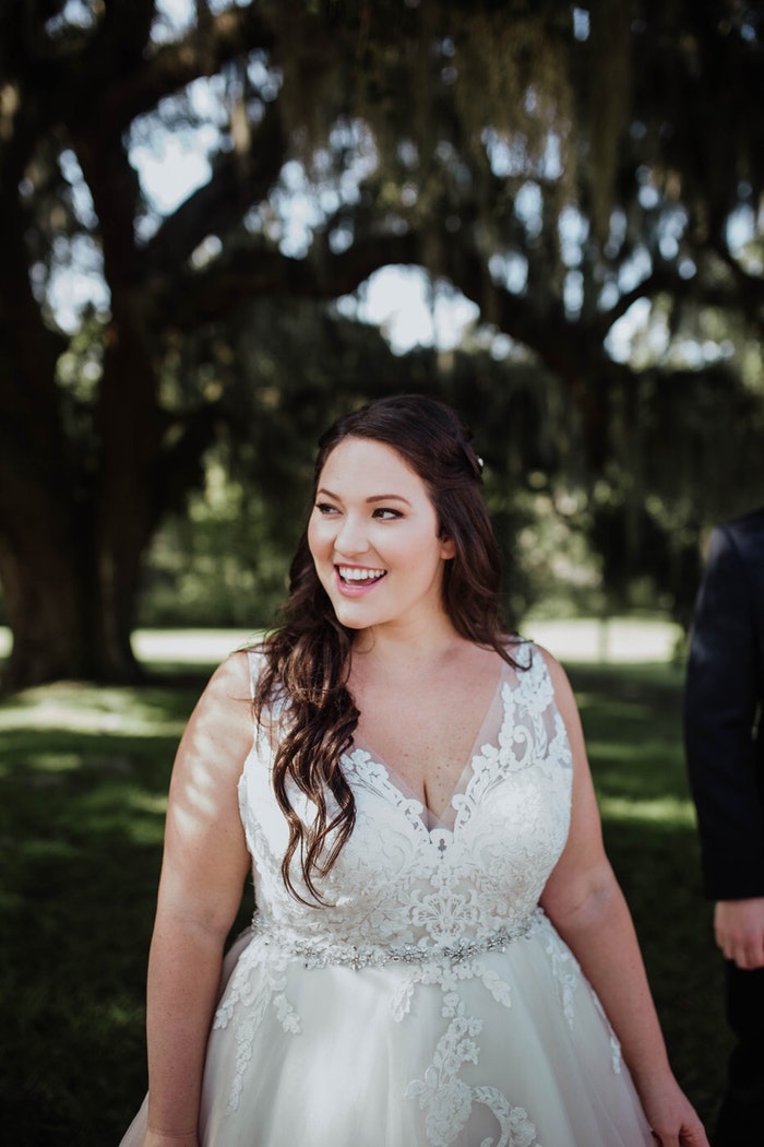 a plus size bride smiles in front of a grove of trees while wearing the sybil dress from maggie sottero designs