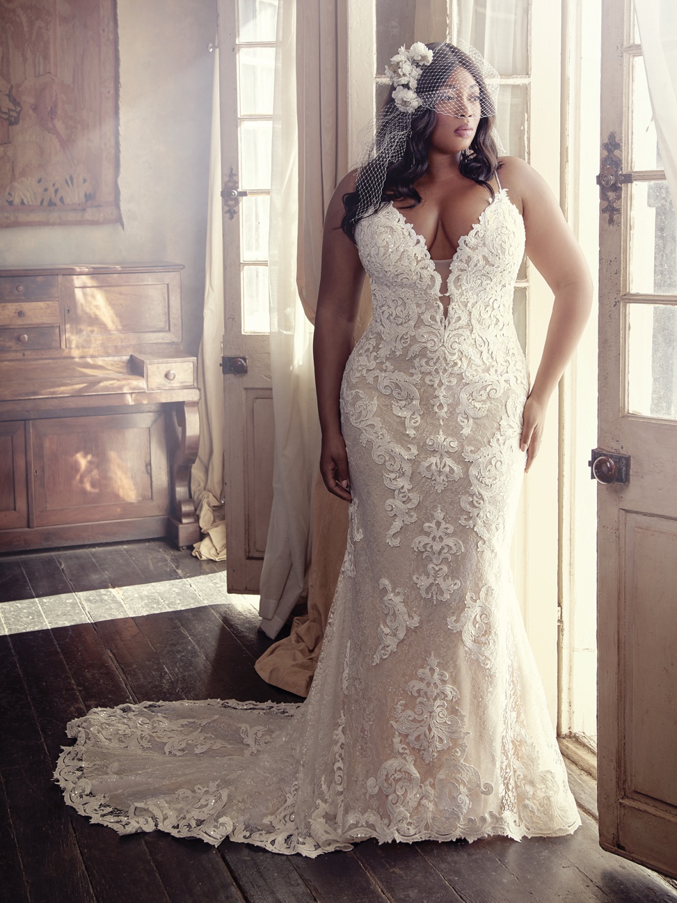 a black plus size bride stands near a set of french doors wearing a pluging lace gown called the tuscany marie from maggie sottero designs with a birdcage veil and faux flowers