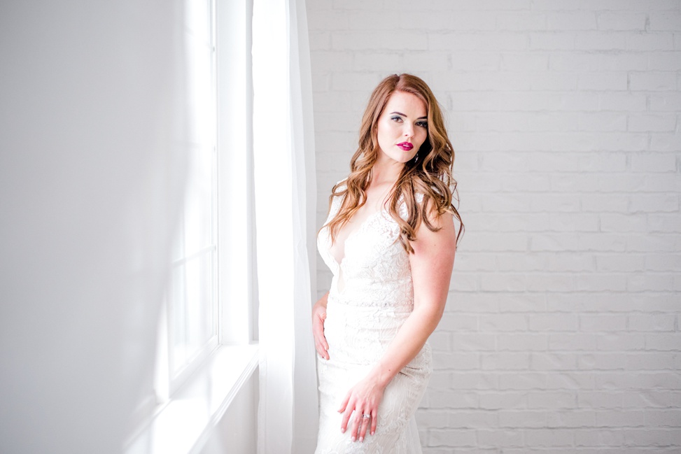 a redhead stands near a window in a bright white room wearing the wyatt dress from sottero and midgley