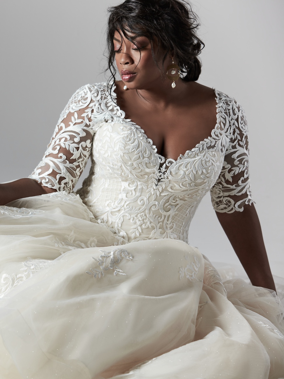 a black bride with a touseled chignon sits posing wearing the river lynette long sleeve lace plus size wedding dress by sottero and midgley