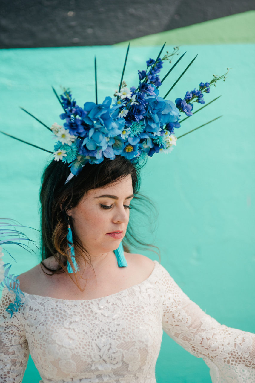A woman wears a madison james long sleeve lace gown holding a blue bouquet and wearing an oversized blue floral headpiece.
