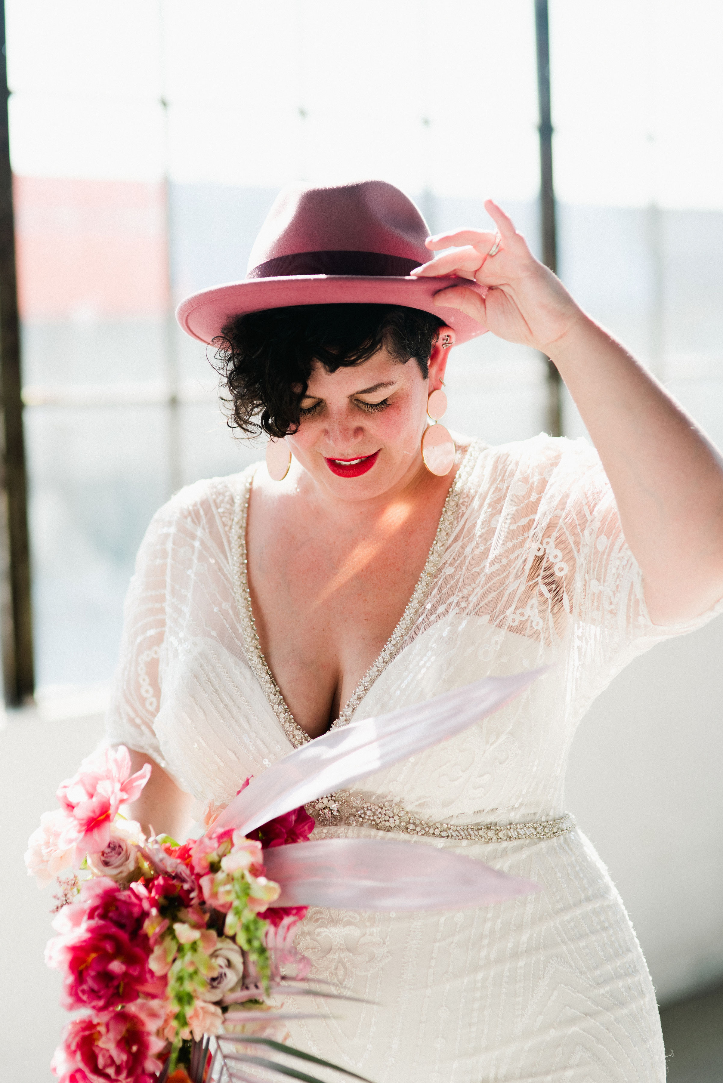 a plus size bride wears a madison james beaded wedding gown holding a pink bouquet with pink earrings, and gently holds a pink felt fedora on her head