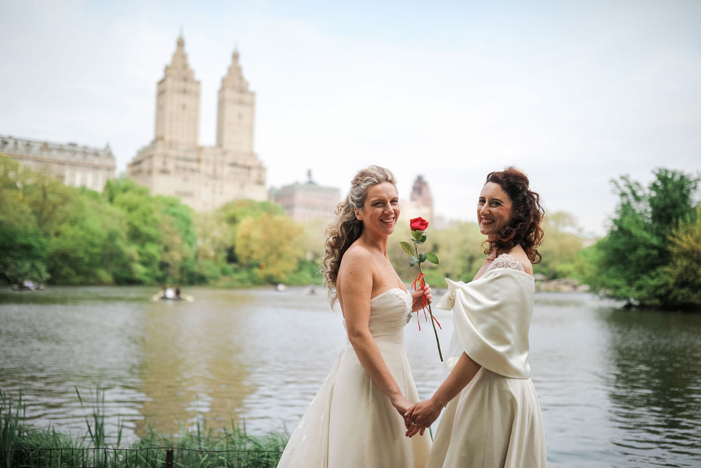 two brides in ivory dresses hold hands and a red rose in Central Park with a lake and the Beresford towers in the background