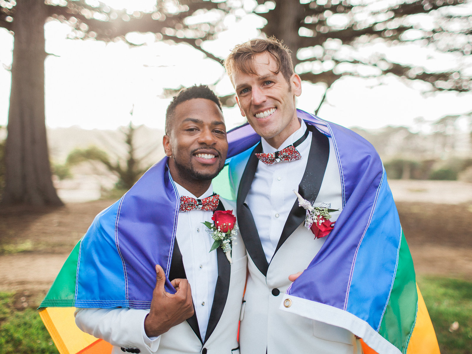 two grooms in white suits stand with their arms around each other under a rainbow flag