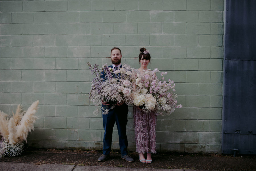 A wedding couple stand against a wall holding large bouquets.