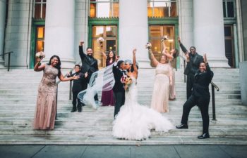 Same-sex black wedding couple, one in black suit and other in a white mermaid ballgroom kiss and fist pump on steps to large stone building while surrounded by their grinning bridal party wearing all kinds of outfits from sequined dresses to suits