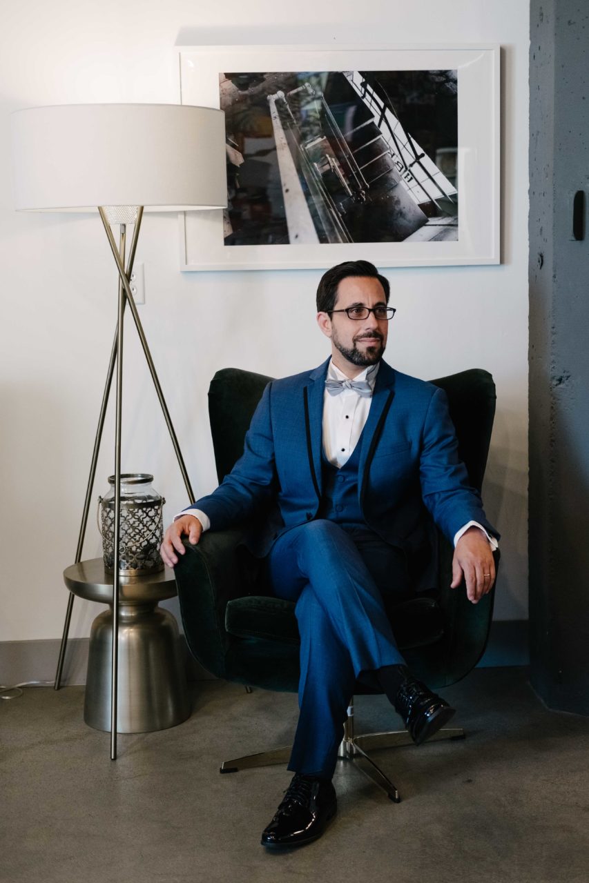a man sits in midcentury velvet chair wearing a mystic blue wedding tuxedo from generation tux