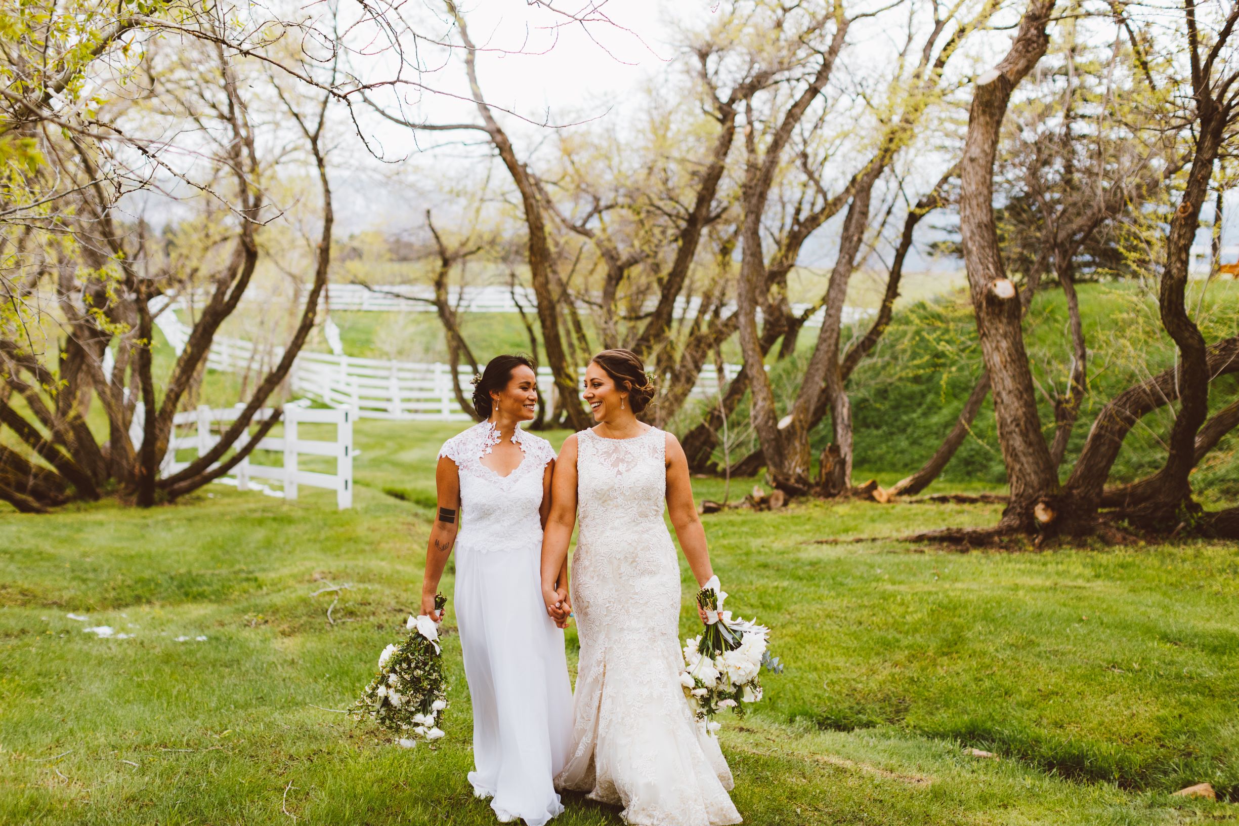 Two women in wedding dresses walk through a field