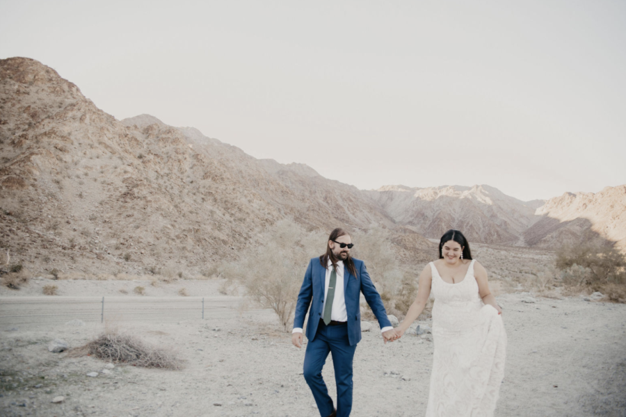 A wedding couple hold hands and walk through a desert
