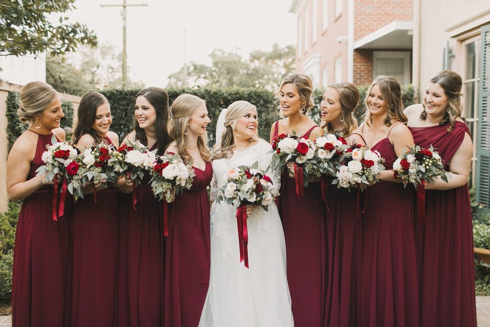 A bridal party stand together wearing deep red bridesmaid dresses