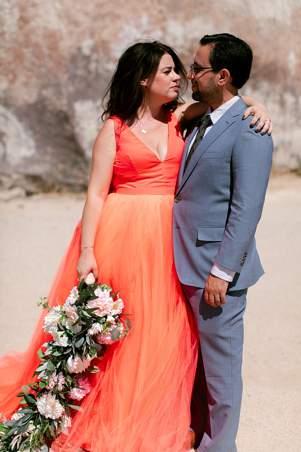 Woman in orange dress, holding a fake wedding flower whip, and man in suit, embrace in the desert