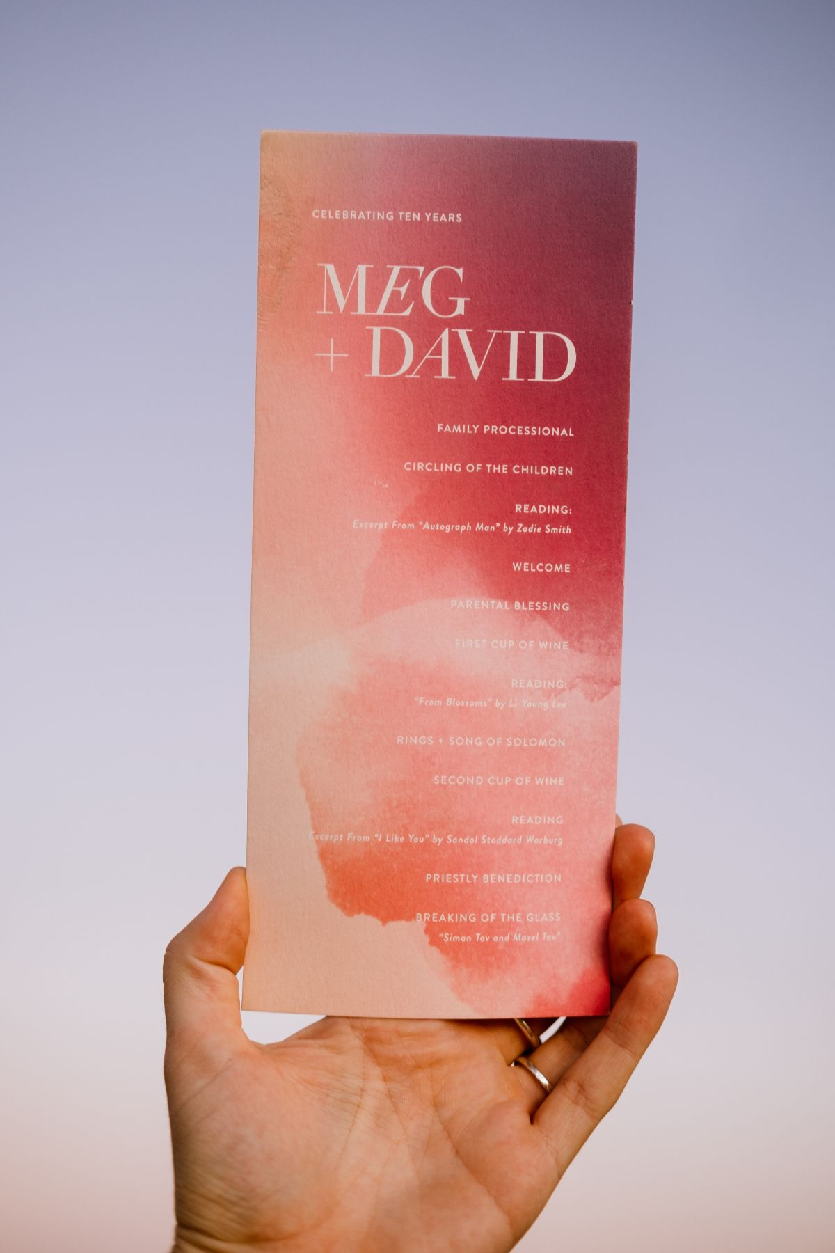 a person's hand holds a pink watercolor wedding program from Minted against a purple sky