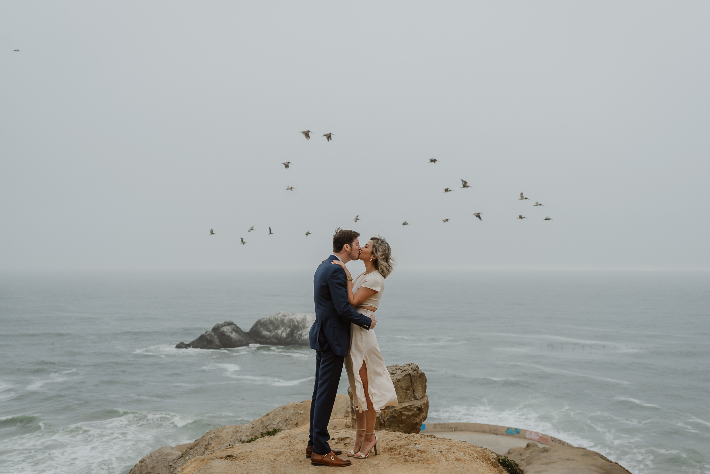 A man and woman kiss on a cliff overlooking the ocean as a flock of birds fly by.