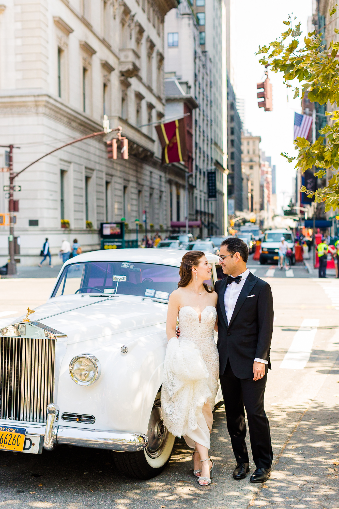 A wedding couple stand next to a vintage car on a busy city street.