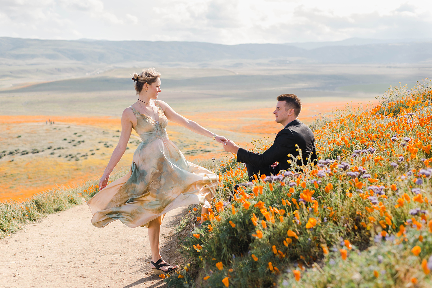 A man sits in a field of wild flowers as a woman holds his hand and stands.