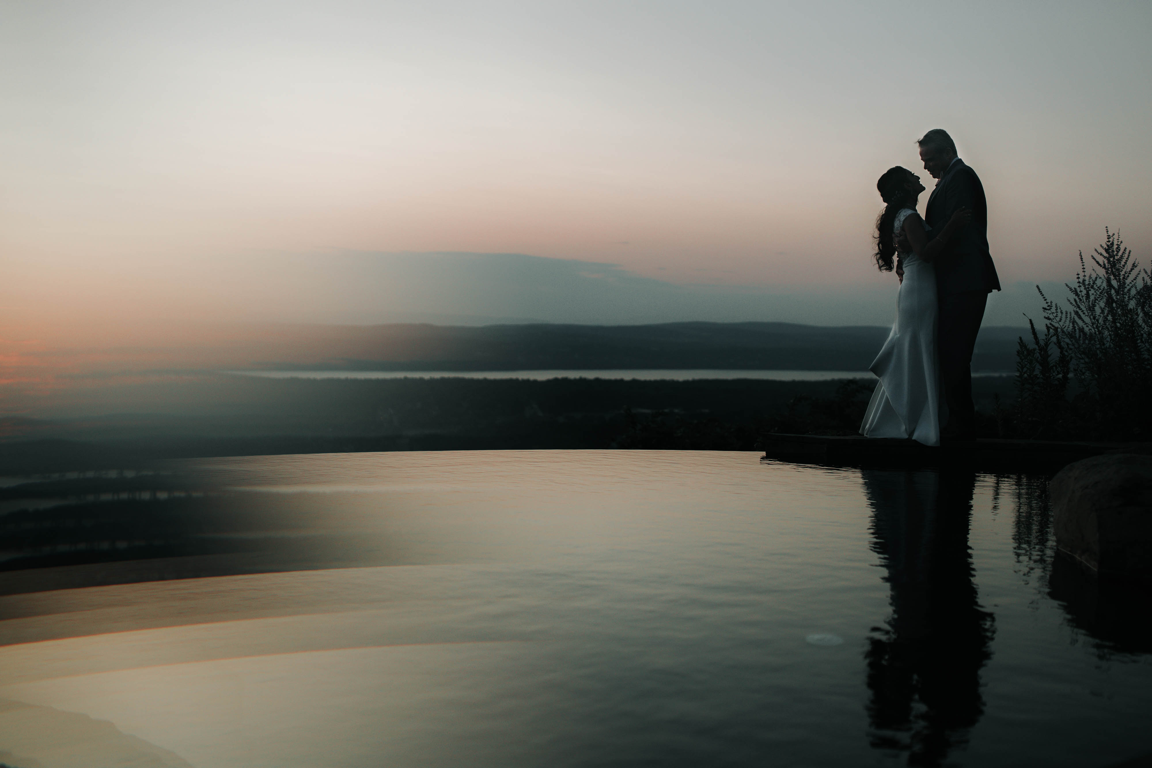 A wedding couple stand and hug at sunset near a reflecting pond.