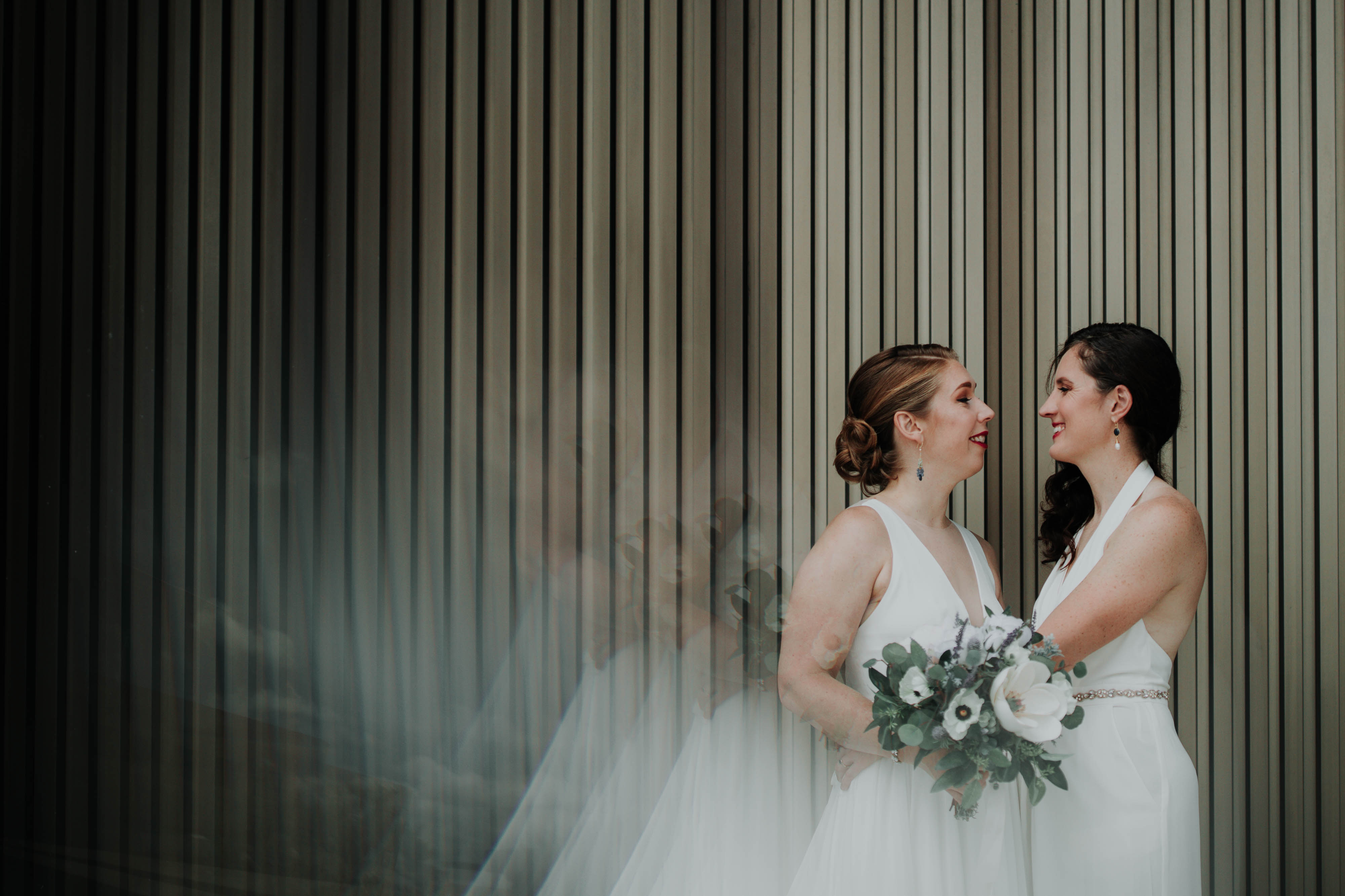 Two women in wedding dresses stand by a wall and embrace.