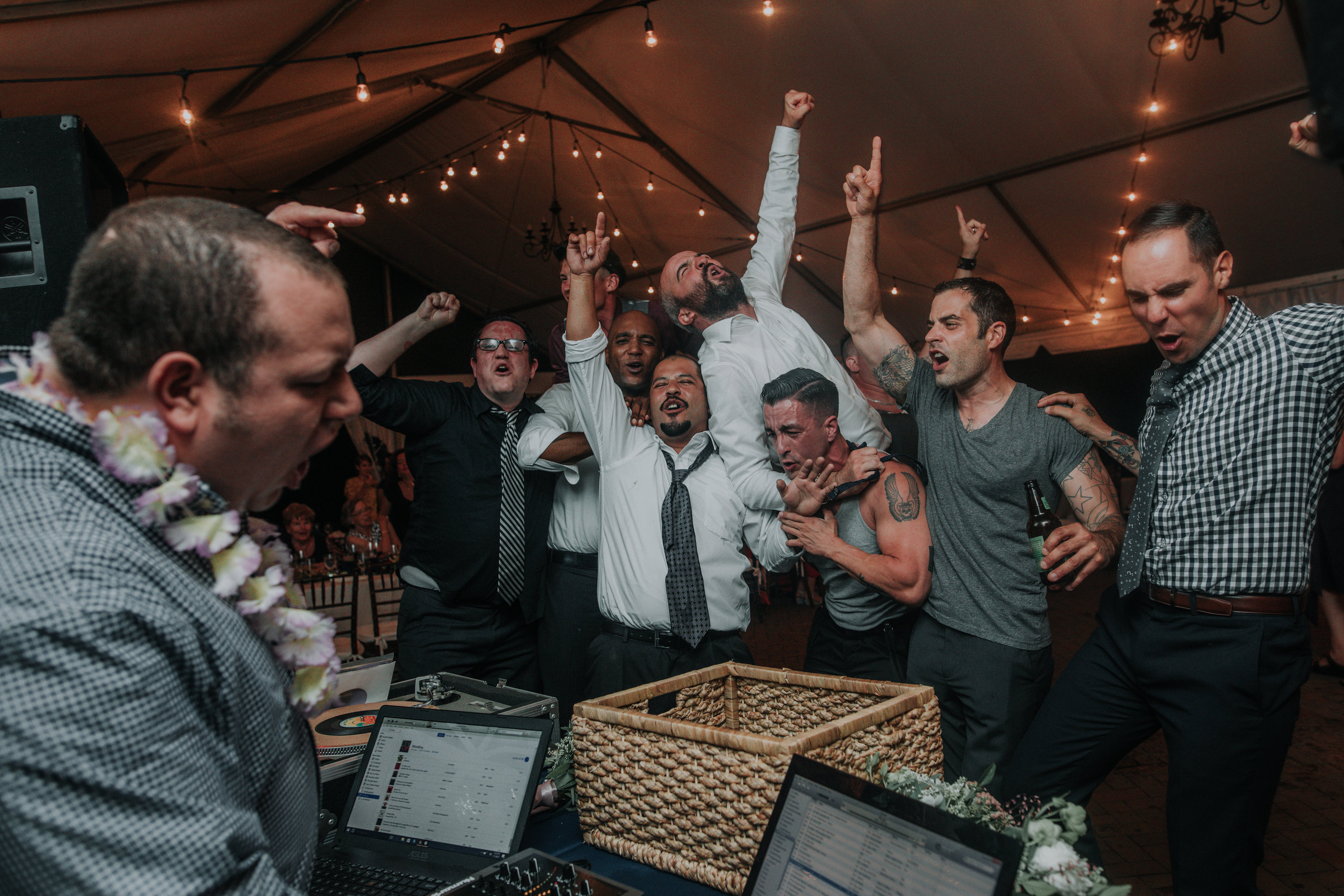  A group of sweaty men dance with 1 finger in the air during a wedding reception.