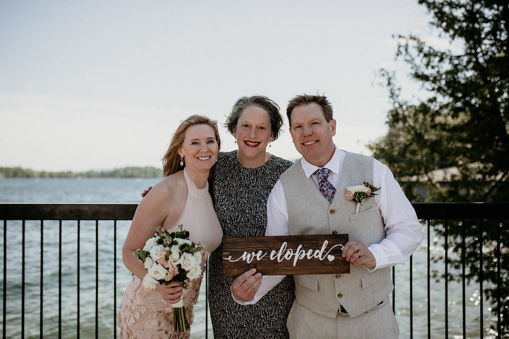 A wedding couple stand with Claudia while holding a sign that says "we eloped"