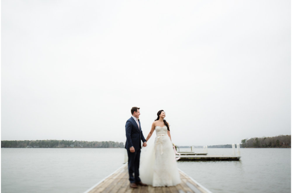 A wedding couple hold hands and stand on a dock.