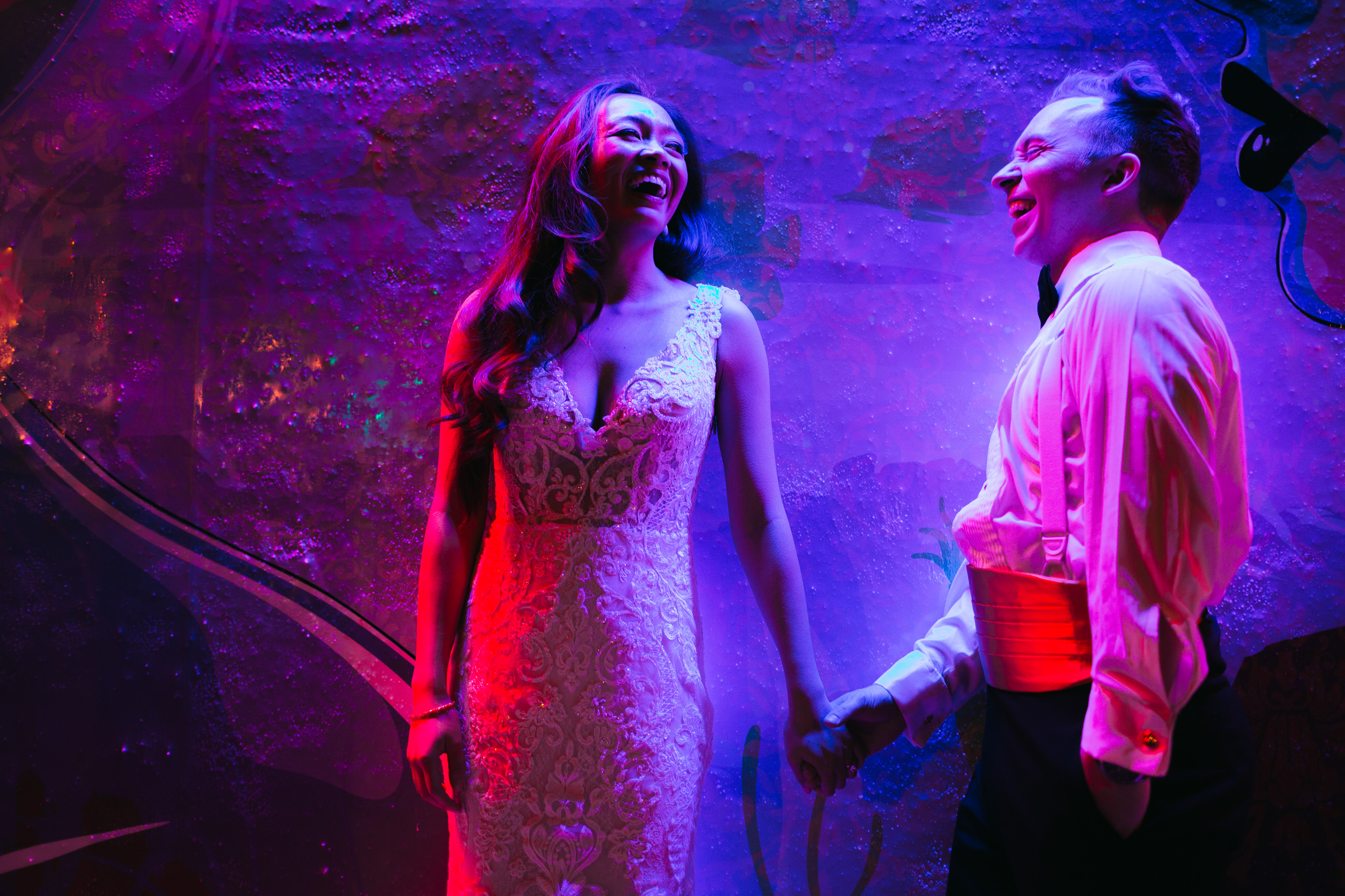 A wedding couple laughs and holds hands under a purple light.