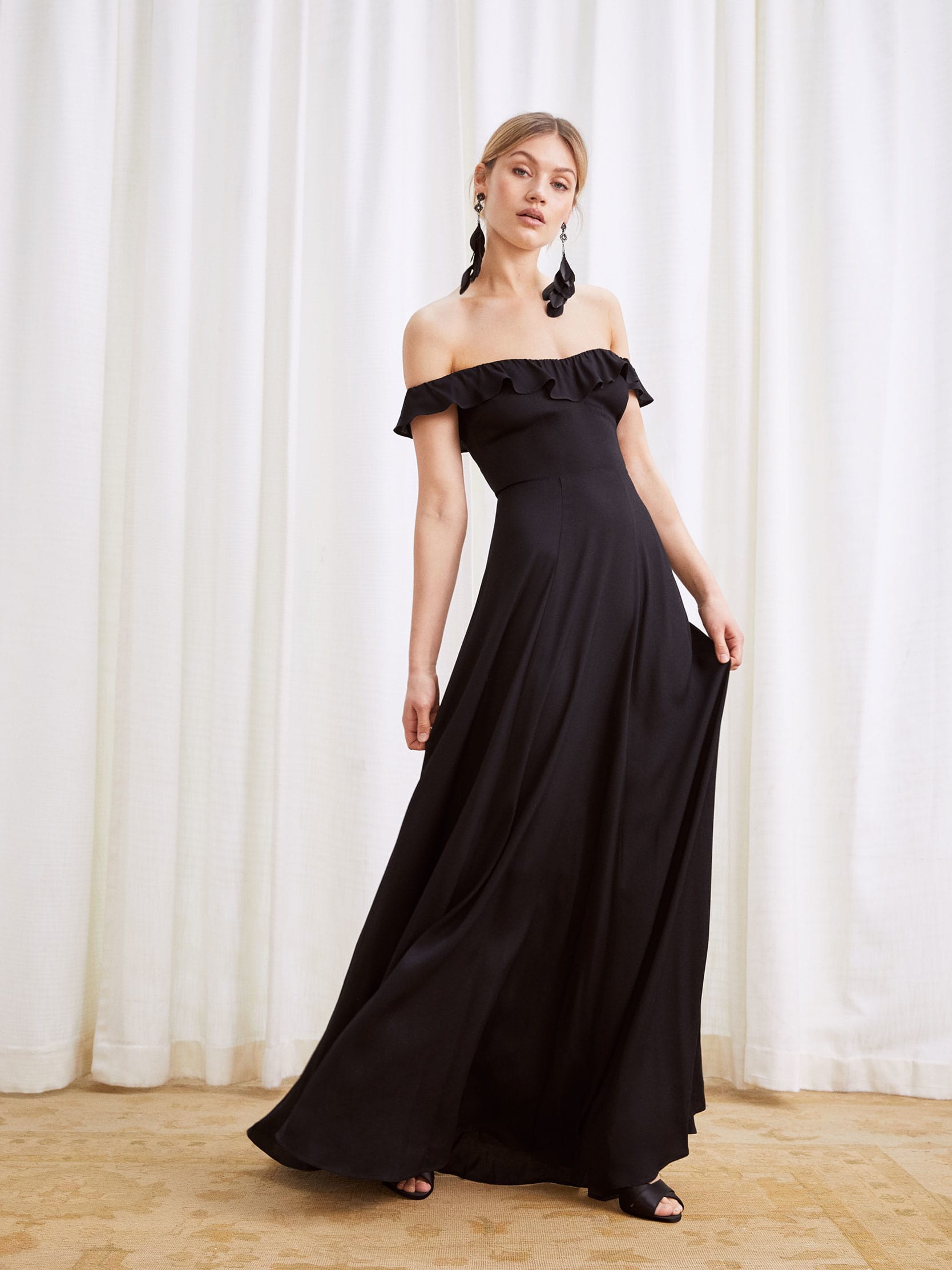 A model wears an off-the-shoulder, floor length dress with a ruffle edged neckline and a center back zipper.