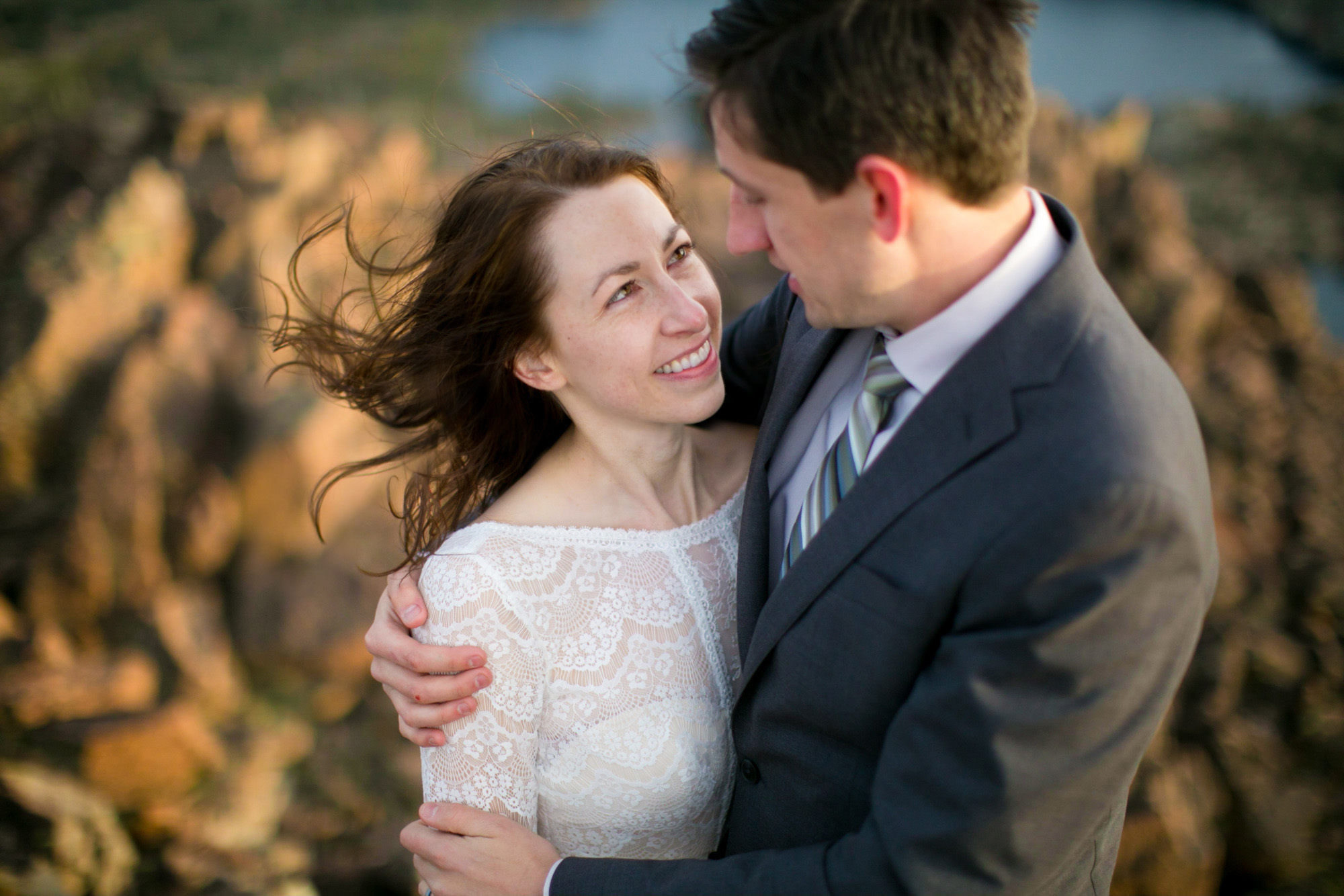 A close-up of bride who smiles up at a groom embracing her as they stand on a cliff.