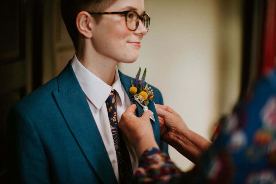 A boutonniere is pinned on a person wearing a blue linen suit and glasses