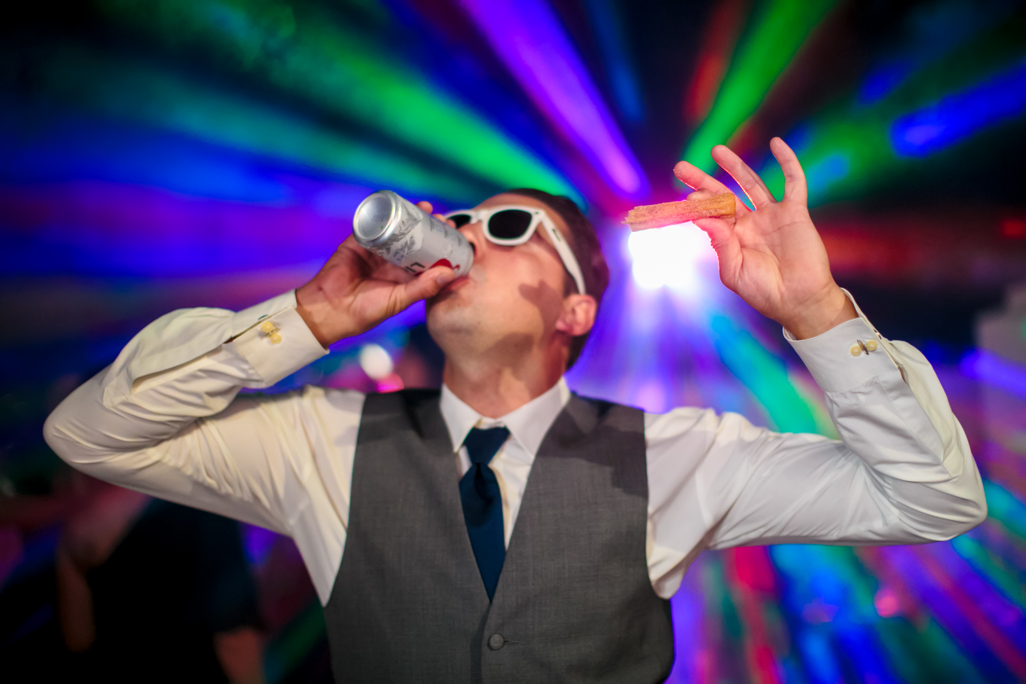 Man dances while drinking a beer and holding a churro in front of a colorful strobe light.