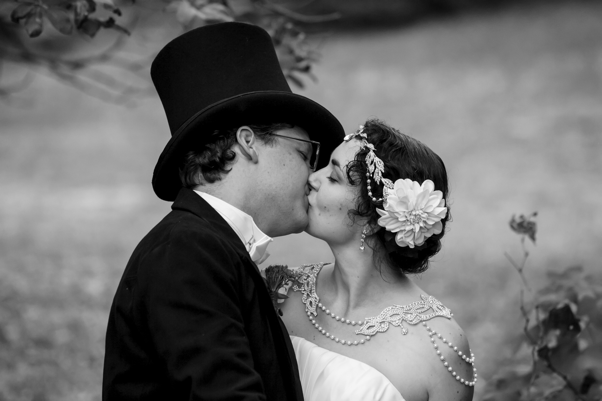 A black and white photo of a wedding couple including a man wearing a top hat and a woman wearing a flower and rhinestone headpiece kissing.