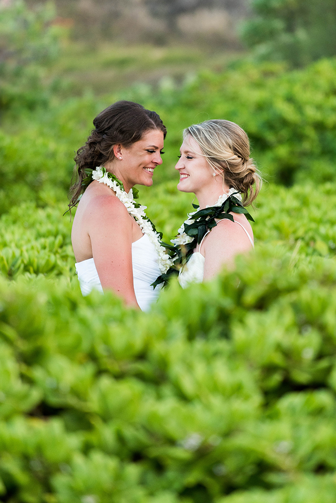 Two brides smile and embrace in a garden.